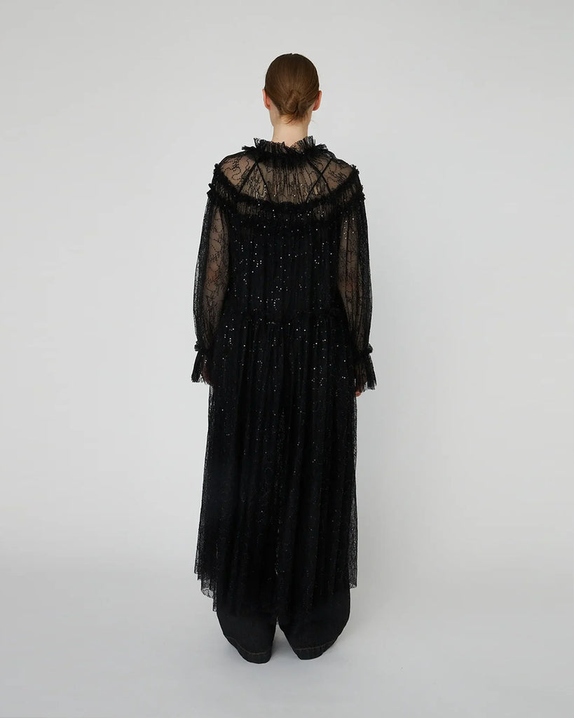 Długa, przezroczysta sukienka Sheer Lace Maxi z koronki z cekinami. Ma zabudowany, efektowny dekolt oraz długie rękawy. Z przodu, na długości ma zapięcia. Można ją nosić zapięta jako sukienkę, bądź rozpiętą jako pelerynę. Sukienka nie posiada podszewki.&nbsp;