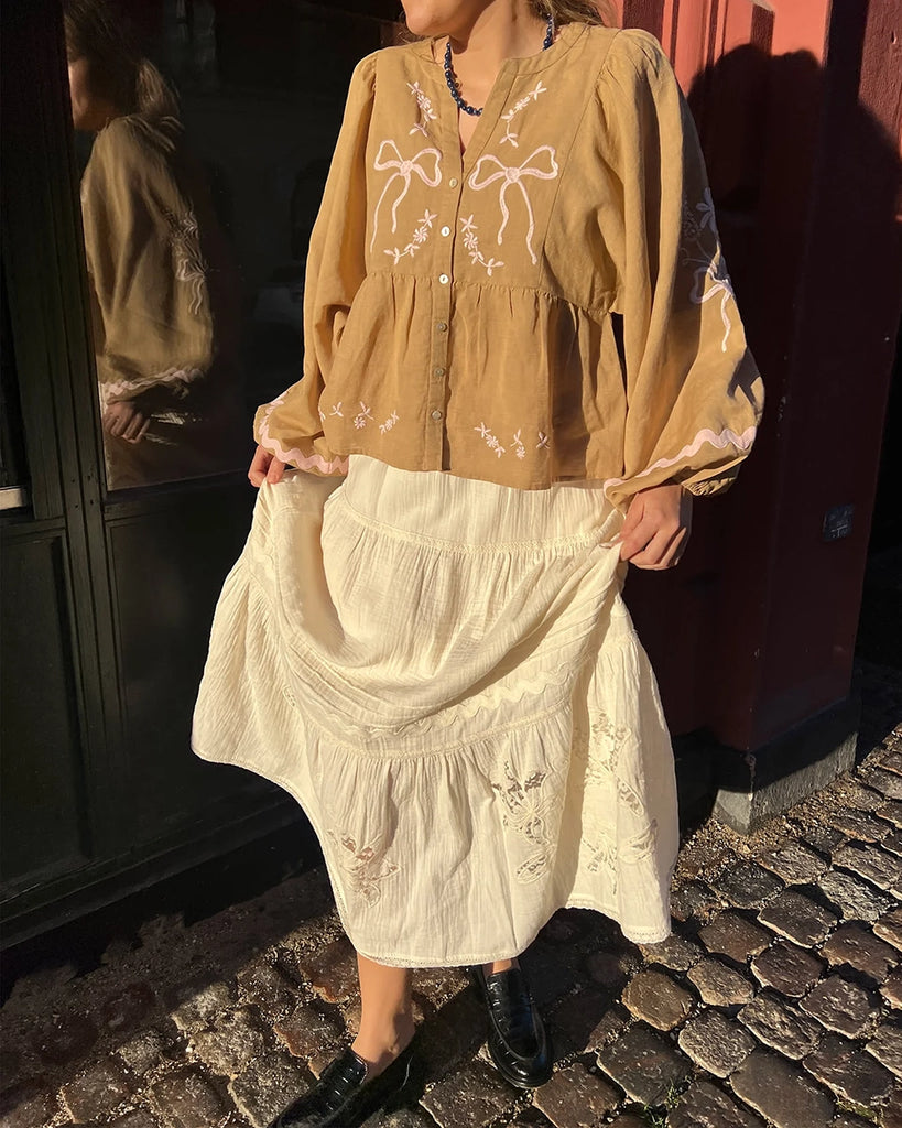 Luźna bluzka Belma SISSEL EDELBO z trójkątnym dekoltem i szerokimi, balonowymi rękawami. Dekorowana pięknym, ręcznie wykonanym haftem z motywami kokard i kwiatów. Odcięta pod biustem. Zapinana na guziki.