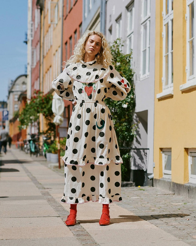 Bawełniana sukienka maxi w grochy Diana SISSEL EDELBO z długimi, balonowymi rękawami, wysokim kołnierzykiem i efektowną falbaną przy dekolcie. Zwężana i elastyczna w talii. U dołu i przy mankietach wykończona falbankami. Dekorowana uroczymi, kolorowymi haftami z motywami serc, kwiatów, gwiazd i księżyca. Obszyta różową lamówką. Sukienka ma długość do kostek