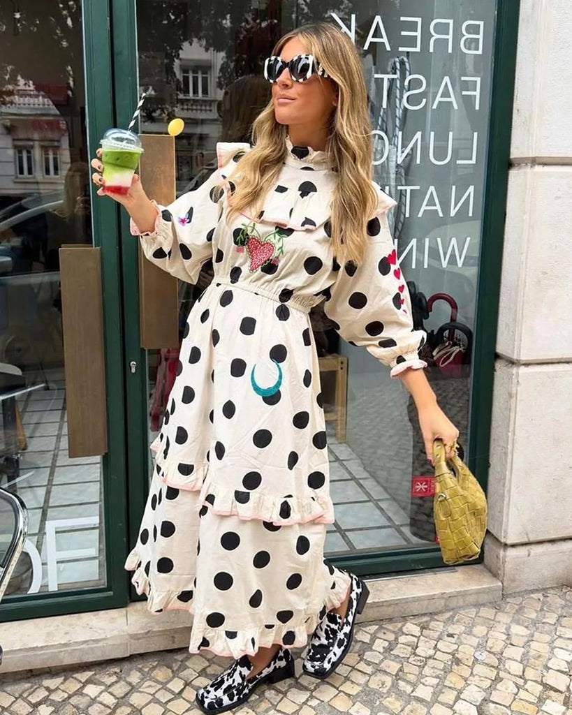 Bawełniana sukienka maxi w grochy Diana SISSEL EDELBO z długimi, balonowymi rękawami, wysokim kołnierzykiem i efektowną falbaną przy dekolcie. Zwężana i elastyczna w talii. U dołu i przy mankietach wykończona falbankami. Dekorowana uroczymi, kolorowymi haftami z motywami serc, kwiatów, gwiazd i księżyca. Obszyta różową lamówką. Sukienka ma długość do kostek