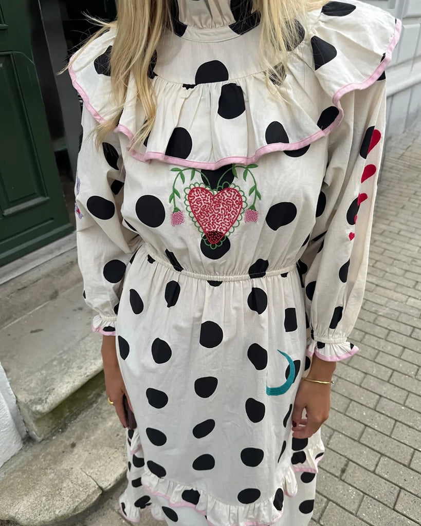 Bawełniana sukienka maxi w grochy Diana SISSEL EDELBO z długimi, balonowymi rękawami, wysokim kołnierzykiem i efektowną falbaną przy dekolcie. Zwężana i elastyczna w talii. U dołu i przy mankietach wykończona falbankami. Dekorowana uroczymi, kolorowymi haftami z motywami serc, kwiatów, gwiazd i księżyca. Obszyta różową lamówką. Sukienka ma długość do kostek