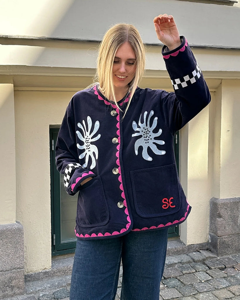 Cienka, krótka, sztruksowa kurtka Maggie SISSEL EDELBO z okrągłym dekoltem i szerokimi rękawami. Dekorowana pięknymi, ręcznie wykonanymi haftami z motywami geometrycznymi, solarnymi i cyrkowymi. Zapinana na guziki. Z przodu posiada dwie, duże kieszenie.