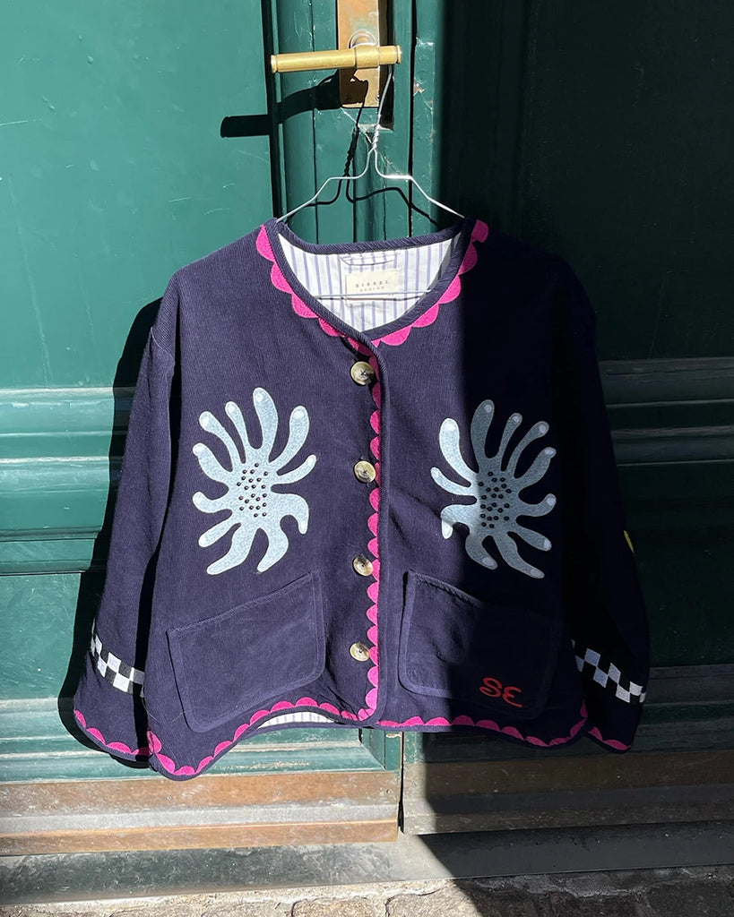 Cienka, krótka, sztruksowa kurtka Maggie SISSEL EDELBO z okrągłym dekoltem i szerokimi rękawami. Dekorowana pięknymi, ręcznie wykonanymi haftami z motywami geometrycznymi, solarnymi i cyrkowymi. Zapinana na guziki. Z przodu posiada dwie, duże kieszenie.