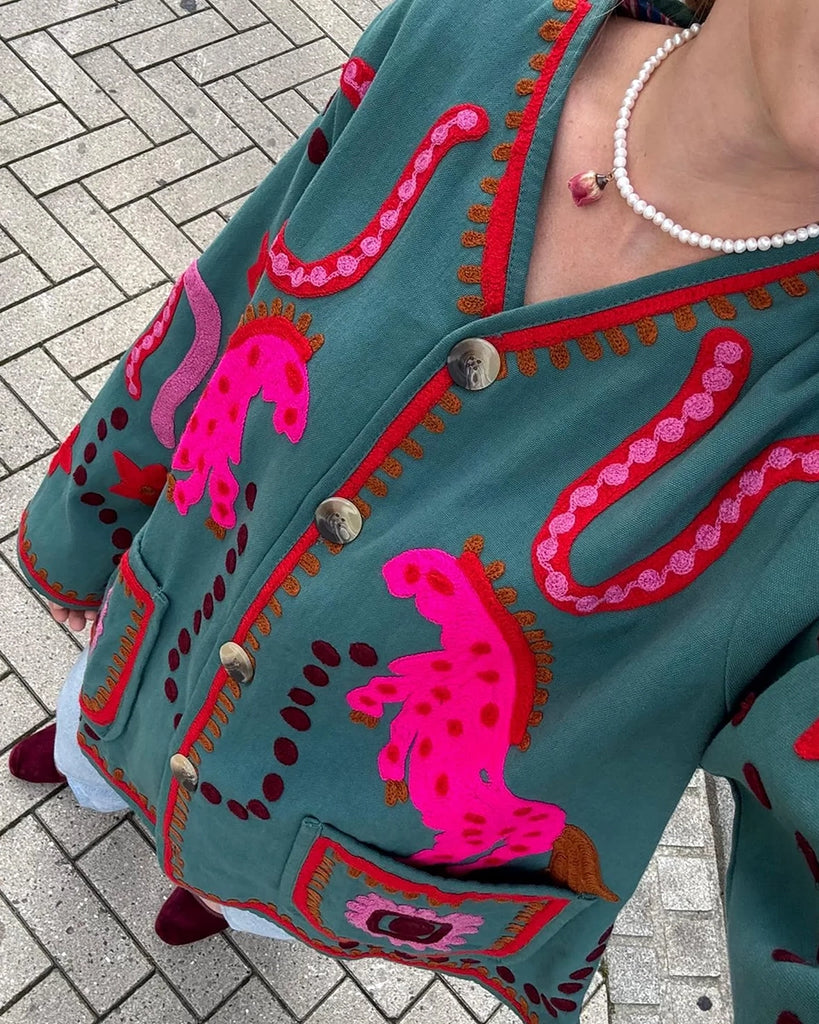 Cienka, krótka kurtka Maggie SISSEL EDELBO z trójkątnym dekoltem i szerokimi rękawami. Dekorowana pięknymi, ręcznie wykonanymi haftami suzani w soczystych, neonowych odcieniach różu . Zapinana na guziki. Z przodu posiada dwie, duże kieszenie.