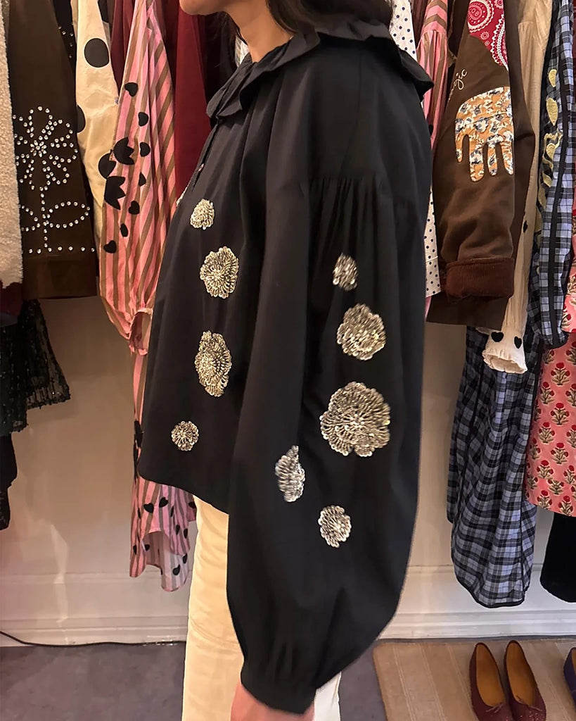 Luźna, bawełniana bluzka Mildred SISSEL EDELBO z okrągłym dekoltem obszytym falbanką, opadającymi ramionami i szerokimi, balonowymi, długimi rękawami. Zapinana na drobne guziki z masy perłowej i elastyczne. Dekorowana efektownymi, kwiatowymi aplikacjami ze srebrnych cekinów i koralików.