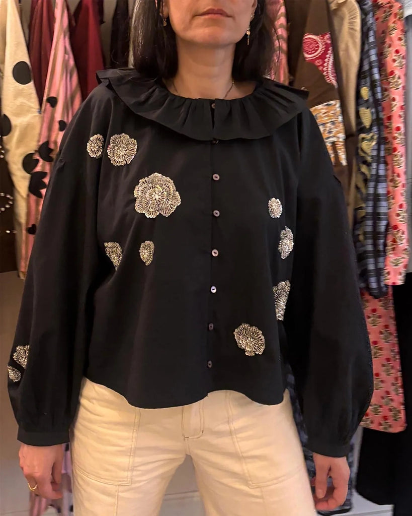 Luźna, bawełniana bluzka Mildred SISSEL EDELBO z okrągłym dekoltem obszytym falbanką, opadającymi ramionami i szerokimi, balonowymi, długimi rękawami. Zapinana na drobne guziki z masy perłowej i elastyczne. Dekorowana efektownymi, kwiatowymi aplikacjami ze srebrnych cekinów i koralików.