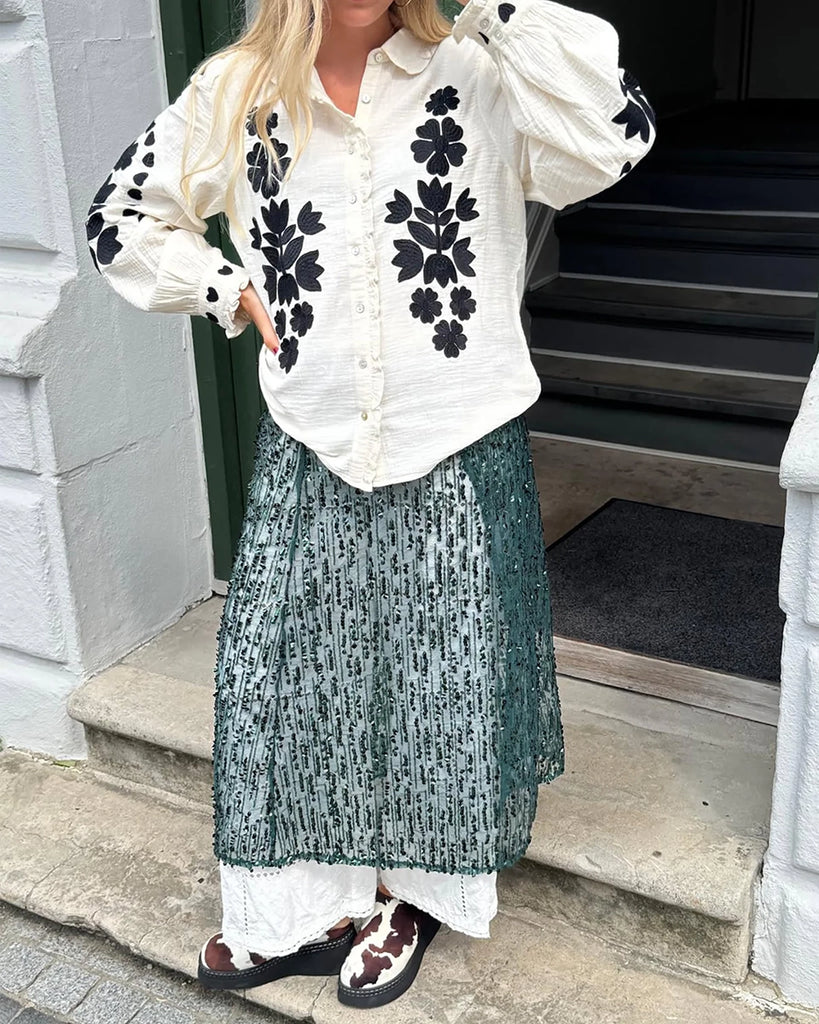 Długa, oversizowa koszula Rae SISSEL EDELBO z falbankowym kołnierzykiem i długimi, balonowymi rękawami wykończonymi mankietami. Zapinana na guziki. Dekorowana kontrastowym, roślinnym haftem