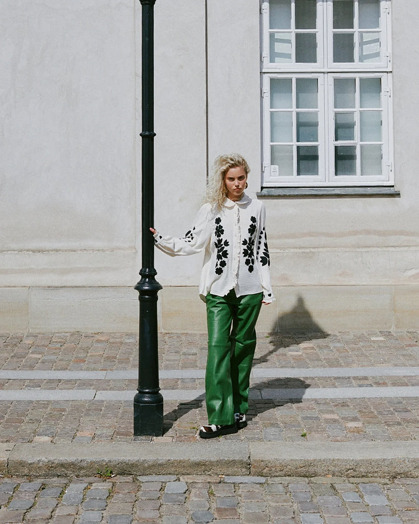 Długa, oversizowa koszula Rae SISSEL EDELBO z falbankowym kołnierzykiem i długimi, balonowymi rękawami wykończonymi mankietami. Zapinana na guziki. Dekorowana kontrastowym, roślinnym haftem