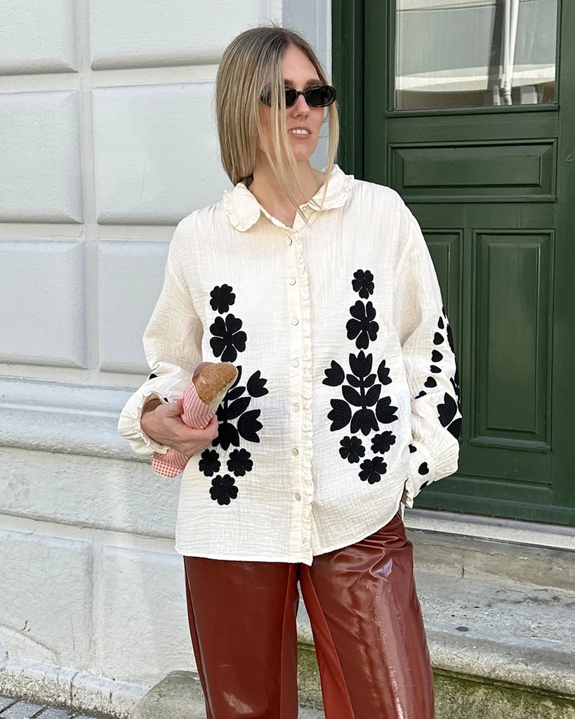 Długa, oversizowa koszula Rae SISSEL EDELBO z falbankowym kołnierzykiem i długimi, balonowymi rękawami wykończonymi mankietami. Zapinana na guziki. Dekorowana kontrastowym, roślinnym haftem