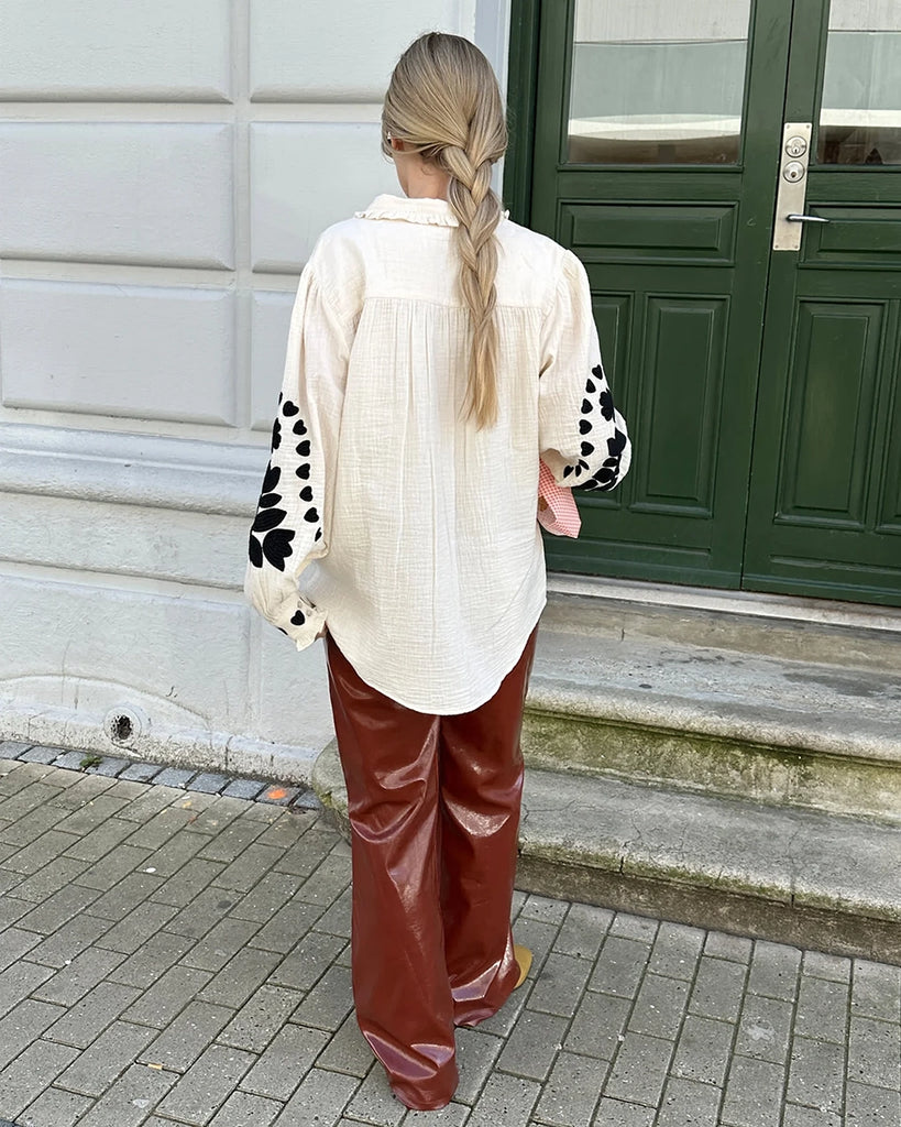 Długa, oversizowa koszula Rae SISSEL EDELBO z falbankowym kołnierzykiem i długimi, balonowymi rękawami wykończonymi mankietami. Zapinana na guziki. Dekorowana kontrastowym, roślinnym haftem