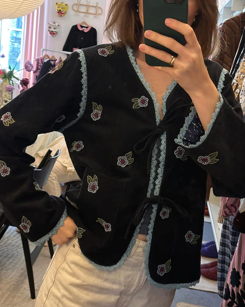 Cienka, krótka, sztruksowa kurtka Tahlia SISSEL EDELBO z trójkątnym dekoltem, wiązana z przodu na kokardki. Wyszywana kolorowymi, drobnymi koralikami w uroczy, kwiatowy wzór. Wykończona drobną, kontrastową koronką.