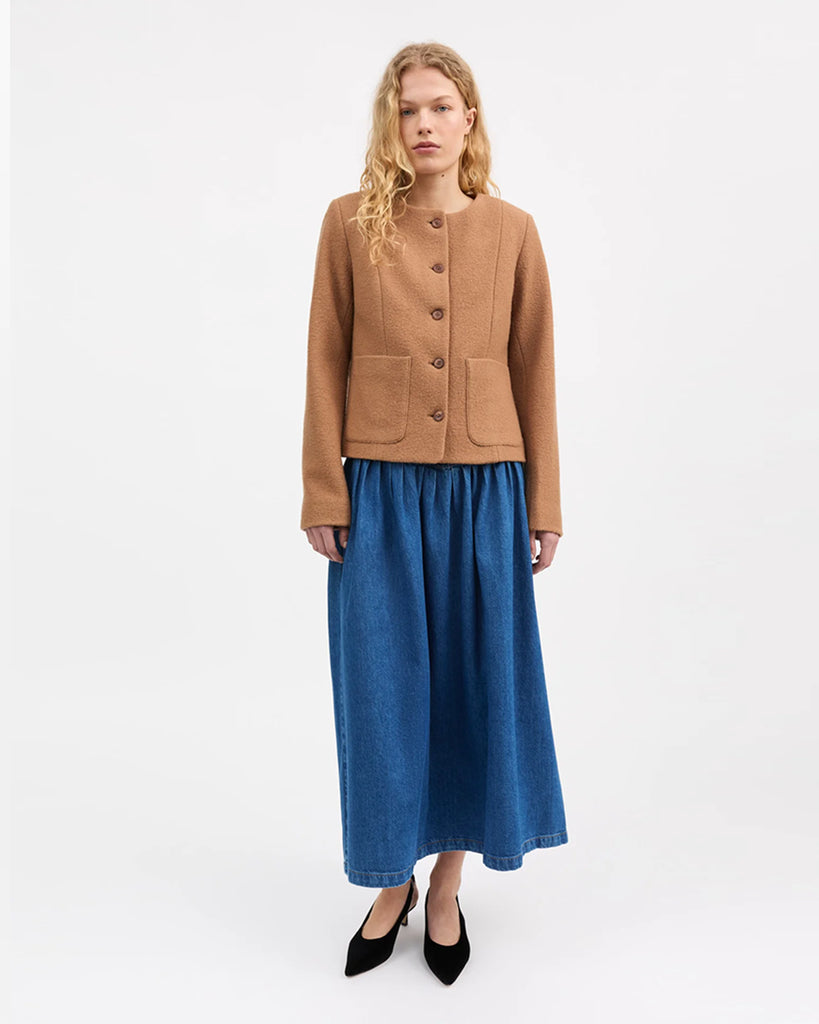 Prosta, lekko pudełkowa kurtka Mona marki SKALL w kolorze karmelowym, z okrągłym dekoltem bez kołnierza, uszyta ze 100% wełny i zapinana z przodu na pięć guzików. Rękawy są długie i proste. Posiada dwie duże kieszenie naszywane z przodu. Podszewka wykonana jest z bawełny organicznej.