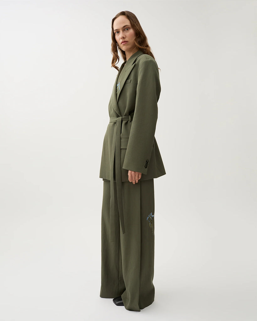 Oversizowa marynarka Stand Dark Moss w kolorze oliwkowej zieleni o wyrazistym, nowoczesnym kroju z haftowanymi zdobieniami w postaci kwiatów. Posiada szerokie klapy w stylu garniturowym oraz pasek w talii, który dodaje kobiecego charakteru. Ma asymetryczne zapięcie, bez widocznych guzików z przodu oraz klasyczne kieszenie z klapką. Zestaw ze spodniami Nora Dark Moss lub z ulubionymi jeansami.