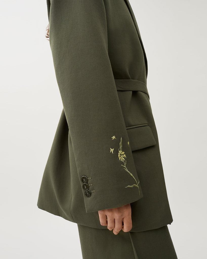 Oversizowa marynarka Stand Dark Moss w kolorze oliwkowej zieleni o wyrazistym, nowoczesnym kroju z haftowanymi zdobieniami w postaci kwiatów. Posiada szerokie klapy w stylu garniturowym oraz pasek w talii, który dodaje kobiecego charakteru. Ma asymetryczne zapięcie, bez widocznych guzików z przodu oraz klasyczne kieszenie z klapką. Zestaw ze spodniami Nora Dark Moss lub z ulubionymi jeansami.
