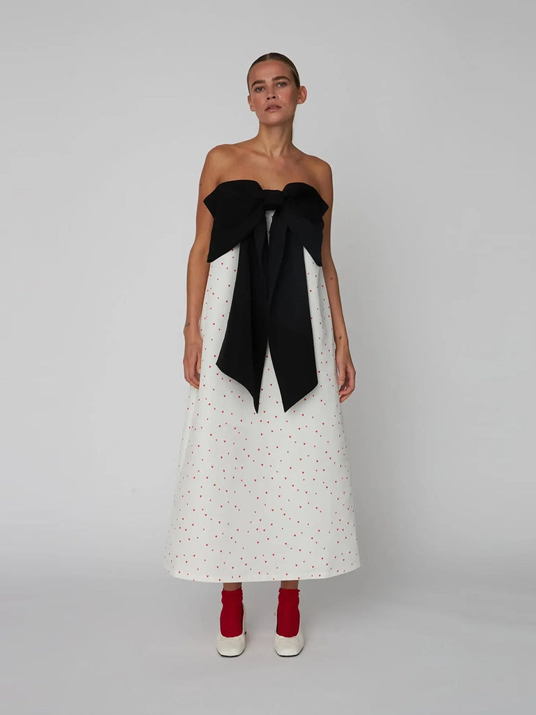 Luźna, wieczorowa sukienka maxi na cienkich ramiączkach Heart Printed Bow STELLA NOVA z dużą, efektowną, czarną kokardą na piersiach. Wykonana ze sztywnej tkaniny w print w drobne serduszka.
