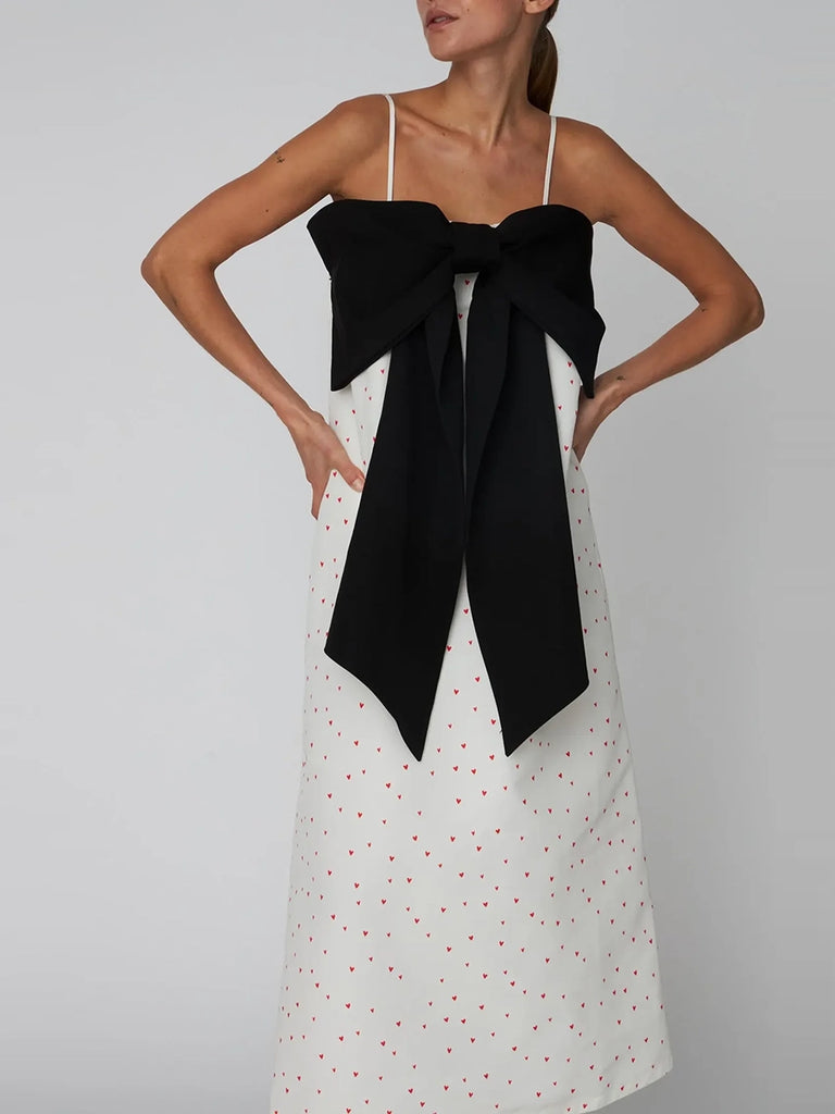 Luźna, wieczorowa sukienka maxi na cienkich ramiączkach Heart Printed Bow STELLA NOVA z dużą, efektowną, czarną kokardą na piersiach. Wykonana ze sztywnej tkaniny w print w drobne serduszka.