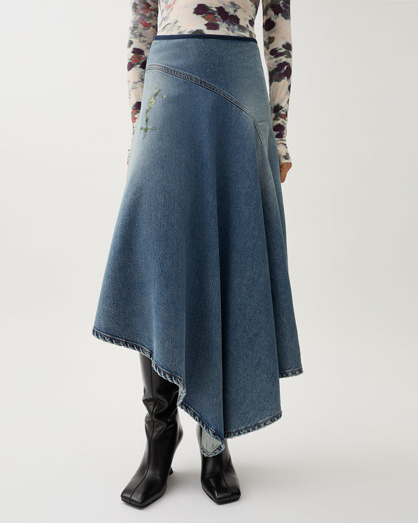 Długa, asymetryczna spódnica Swoosh Indigo z wysokim stanem, w niebieskim odcieniu denimu z przetarciami w stylu vintage i kwiatowymi haftami. U góry ma granatową lamówkę i zapięcie na suwak z boku.&nbsp;