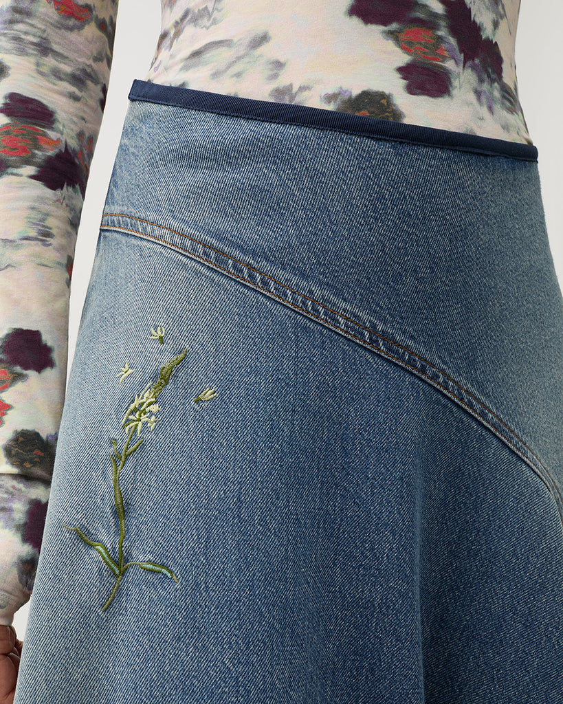 Długa, asymetryczna spódnica Swoosh Indigo z wysokim stanem, w niebieskim odcieniu denimu z przetarciami w stylu vintage i kwiatowymi haftami. U góry ma granatową lamówkę i zapięcie na suwak z boku.&nbsp;