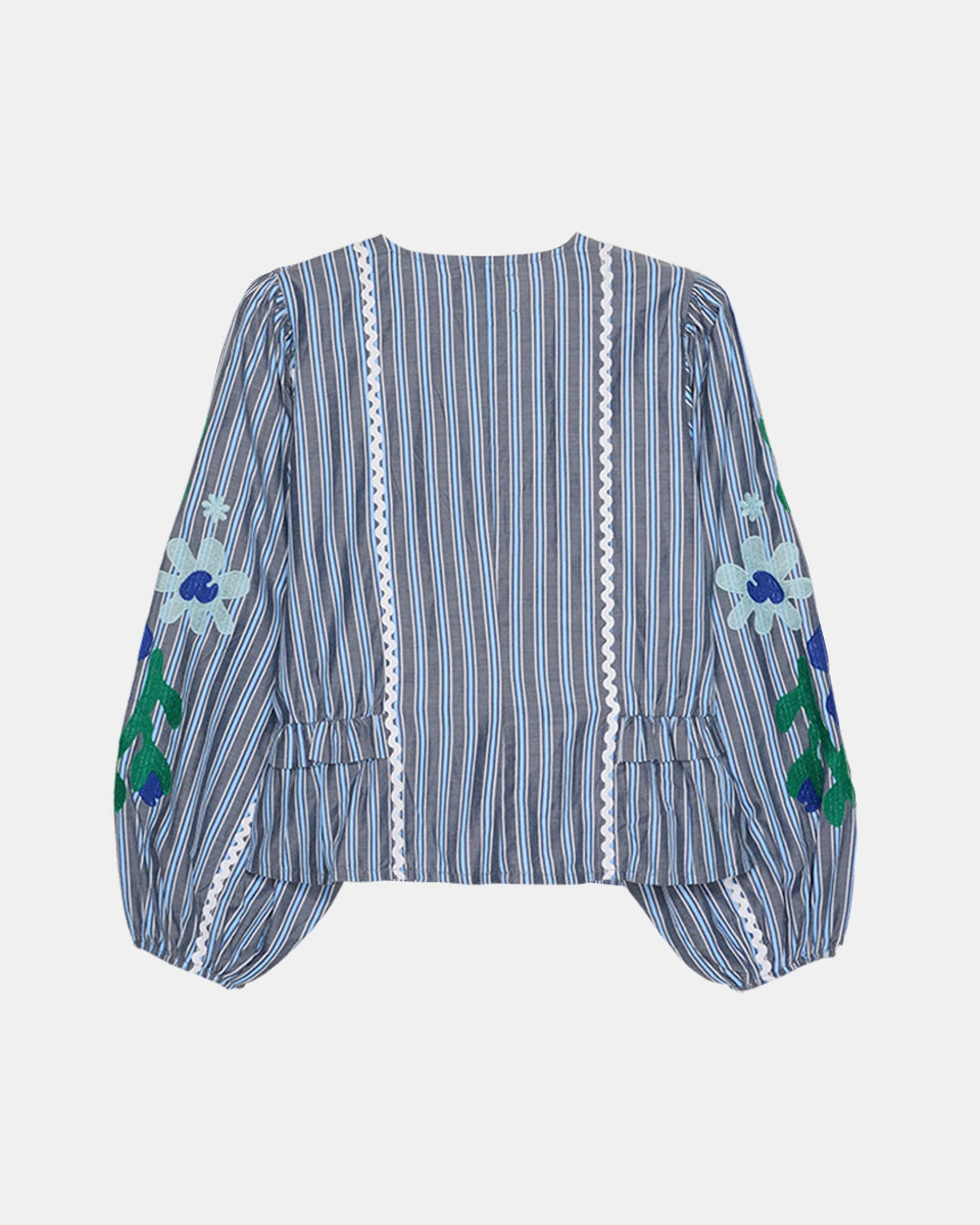 Luźna, koszulowa bluzka Veda Grey Stripes marki SISSEL EDELBO z miękkiej bawełny w szare paski. Ma głęboki dekolt w serek i zapinana jest na ciemnie guziki z masy perłowej z elastycznymi pętelkami. Koszula ma talię z drobnymi falbankami i wstawkami po bokach oraz panele biegnące wzdłuż przodu. Szerokie, bufiaste rękawy zdobi duży wyrazisty haft kwiatowy i zygzakowata bawełniana taśma. Otwory na rękawy posiadają gumkę wewnątrz, co zapewnia wygodne dopasowanie. Uszyta została z bawełnianych resztek tkanin.&nb