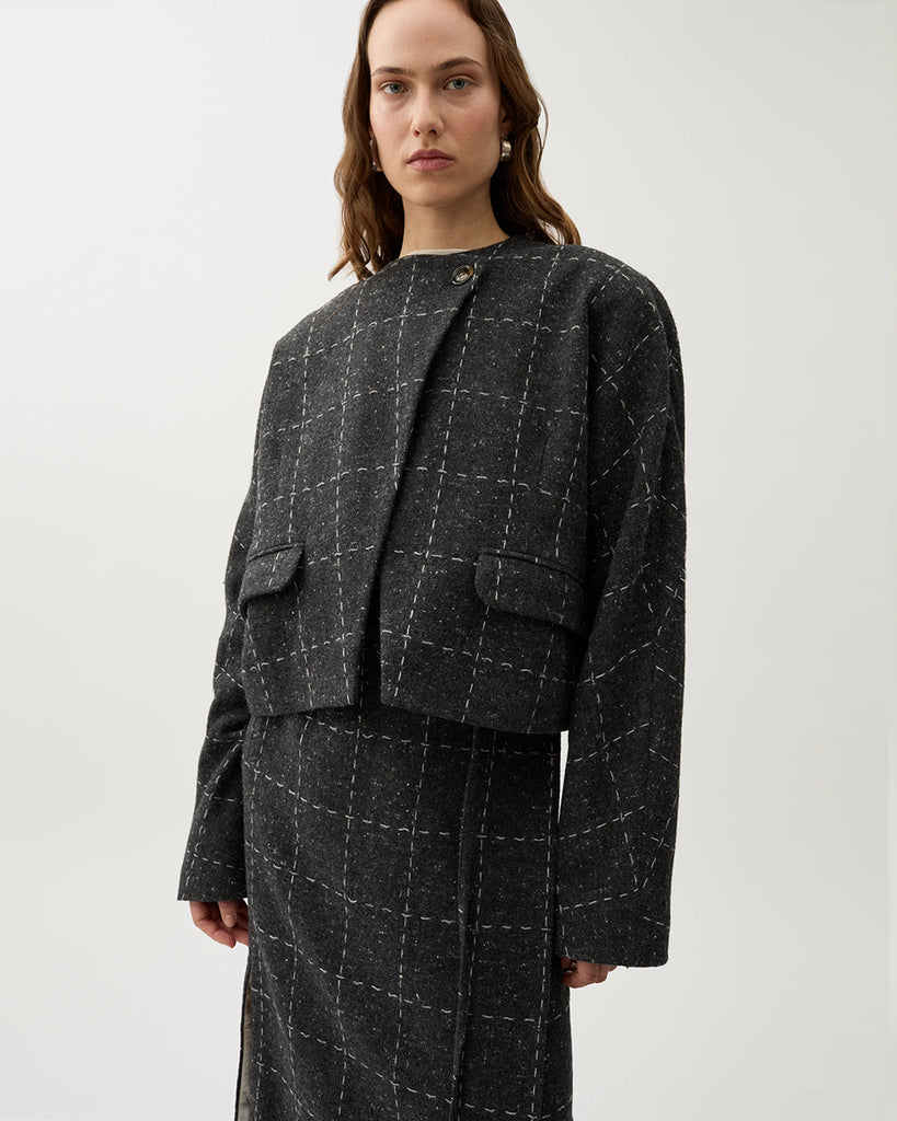 Krótka, luźna marynarka Zola Check Grey Melange w kolorze ciemnoszarym w kratkę z szerokimi rękawami i okrągłym dekoltem. Jest zapinana na jeden guzik pod szyją i posiada kieszenie boczne. Wykonana z mieszanki wełny. Zestaw ją z spódnicą Blanket Check lub ulubionymi jeansami.&nbsp;